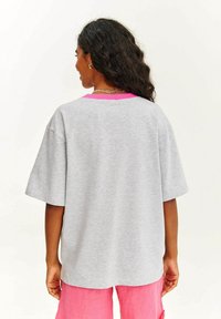 Šedé oversized tričko s růžovým límečkem, krátkými rukávy a volným střihem. Materiál vypadá měkce s hladkým povrchem.