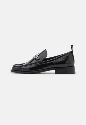 Mocasín de charol negro con tacón bajo, adorno de hebilla metálica plateada, costura tipo mocasín en la puntera y lengüeta trasera para facilitar el calzado.