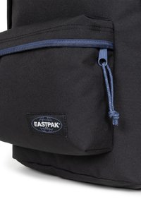 Černý látkový batoh s přední kapsou, která má modrý zip a šňůrku. Logo Eastpak je viditelné na boku. Texturovaný povrch.
