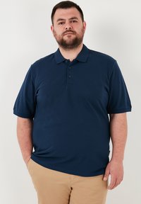 Tamsiai mėlynas polo marškinėliai, pagaminti iš lygios medžiagos, su dviem mygtukais ir trumpomis rankovėmis. Nešiojami su kreminėmis kelnėmis.
