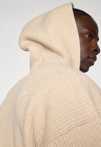 Felpa beige a coste con ampio cappuccio e un sottile dettaglio del logo. Il tessuto testurizzato aumenta il calore e il comfort.