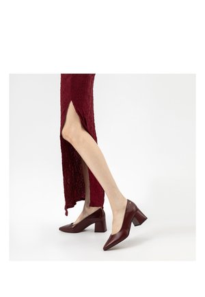 Frau trägt ein strukturiertes burgunderrotes Kleid mit seitlichem Schlitz und passende burgunderrote Blockabsatzschuhe, steht auf einer weißen Oberfläche.