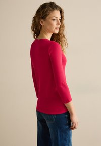 Haut ajusté à manches longues en coton rose vif. Texture lisse avec un col rond. Associé à un pantalon en denim présentant des poches arrière.