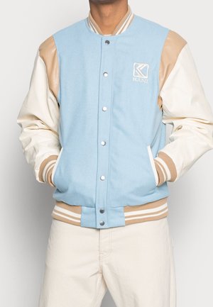 La personne porte une veste universitaire bleu clair et beige avec des manches blanches, des poignets beige rayés et des poches avant avec les mains à l'intérieur.