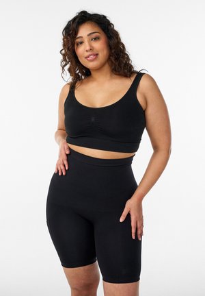 Woman with curly hair wearing a black cropped tank top and high-waisted biker shorts, standing against a plain white background.