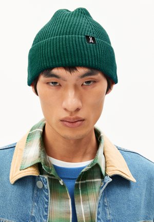 Green ribbed beanie featuring a pointed top and a small black tag with a logo. Worn with a denim jacket and plaid shirt underneath.