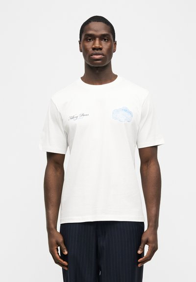 Άνδρας που φοράει λευκό κοντομάνικο μπλουζάκι με μπλε κείμενο και μπλε γραφικό νεφοπλέγμα σε απλό λευκό φόντο.