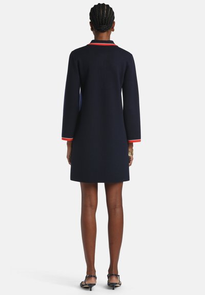 Žena s vrkočmi, oblečená v námornícky modrých šatách s dlhým rukávom a červeným lemovaním na golieri a manžetách, stojaca otočená chrbtom, čierne topánky s nízkym opätkom.