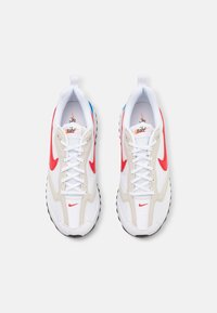 Baskets blanches et beige avec des accents rouges et un onglet de talon bleu. Caractérisées par un mesh texturé et une semelle noire épaisse avec un motif de crantage.