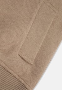 Beige ullblandad sweatshirt med en texturerad yta, ribbade muddar och en synlig sidoficka för funktionalitet.