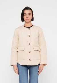 Μπεζ μπουφάν με καπιτονέ σχέδιο, στρογγυλή λαιμόκοψη, πέντε χρυσά κουμπιά και δύο μπροστινές τσέπες. Το ύφασμα έχει λεία υφή, με μαύρες λεπτομέρειες.