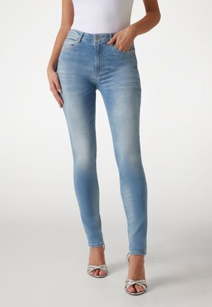 Kobieta w jasnoniebieskich dopasowanych dżinsach i srebrnych butach na wysokim obcasie z odkrytymi palcami, pozująca z jedną ręką w kieszeni na białym tle.