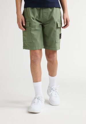 Pantaloncini cargo verdi con due grandi tasche laterali, realizzati in materiale leggero. Indossati con sneakers bianche e calzini bianchi a lunghezza media.