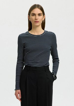 Jeune femme aux cheveux bruns raides portant un haut à manches longues rayé marine et blanc et un pantalon noir taille haute, debout avec une main dans la poche.
