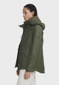 Veste imperméable vert olive avec capuche, fermeture éclair à l'avant, détails à boutons-pression et patch logo sur la manche gauche.