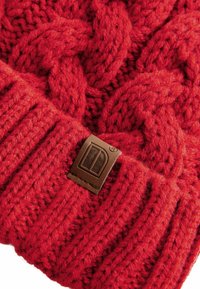 Bonnet tricoté rouge présentant un motif de câble texturé, une bande côtelée et un patch en cuir marron avec un détail de logo.