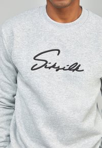 Felpa grigia in materiale morbido, con un logo nero ricamato in evidenza, "Sitsilk", in carattere corsivo sul davanti.