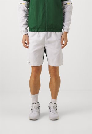 Pantaloncini sportivi bianchi con accenti laterali verdi, abbinati a una giacca con zip verde e bianca. Il modello indossa scarpe sportive bianche e calzini.