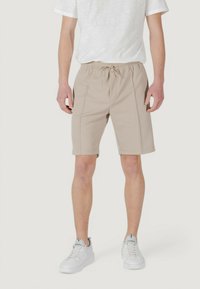 I pantaloni corti beige con vita elastica presentano tasche laterali e dettagli cuciti, abbinati a una maglietta grigio chiaro e sneaker bianche.