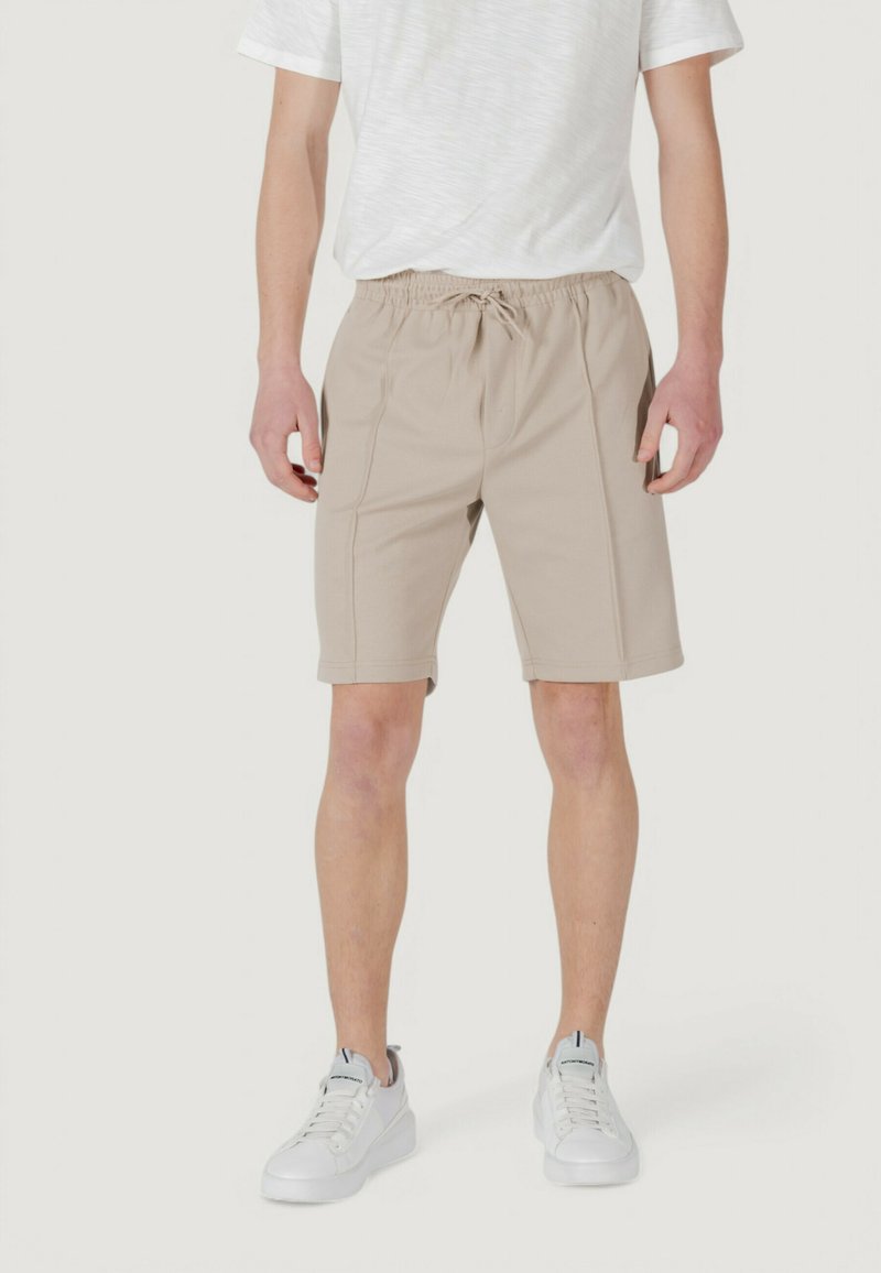 I pantaloni corti beige con vita elastica presentano tasche laterali e dettagli cuciti, abbinati a una maglietta grigio chiaro e sneaker bianche.