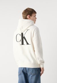 Sudadera crema con un gran logo negro "CK" en la espalda. Fabricada con un tejido suave, cuenta con capucha con cordón y puños acanalados.