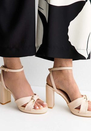 Pieds portant des sandales beiges à talons blocs avec des brides à la cheville, assorties à une jupe midi à motif noir et blanc.