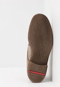 Chausson marron avec une semelle texturée, présentant un dessus doux et un accent rouge sur le talon. Le design est simple et fonctionnel.