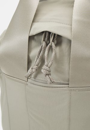 Sac à dos beige avec double poignée de transport, fermeture par cordon et un logo patch carré sur le bas à droite à l'avant.