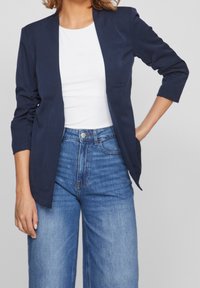 Námořnický blazer s fitted siluetou a dlouhými rukávy, nošený přes bílé tričko, spárovaný s džínami s vysokým pasem v modré barvě. Hladká textura látky.