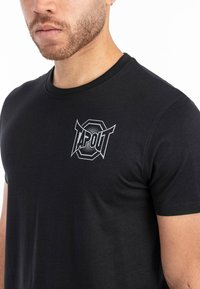 Must have musta T-särk mustast puuvillast, ümmarguse kaelusega ja lühikeste varrukatega. Vasakul rinnal on hõbedane "TAPOUT" logo. Sile tekstuur.