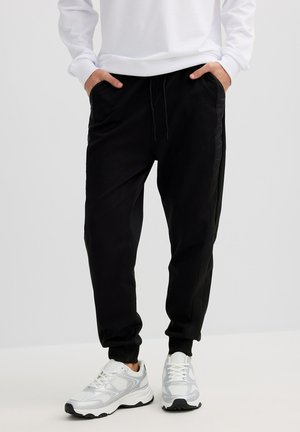 Persona vistiendo pantalones jogger negros con bolsillos laterales y zapatillas deportivas blancas, de pie con las manos en los bolsillos sobre un fondo llano.