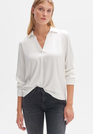 White, long-sleeve blouse with a V-neck and buttoned cuffs, made of smooth fabric. Paired with dark jeans, highlighting a relaxed fit.