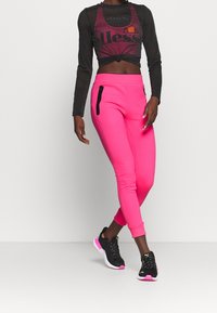 Zwarte lange mouwen top met een geknoopte voorkant, en aansluitende roze joggers met zwarte ritszakken. Zwarte en roze sneakers maken de look compleet.