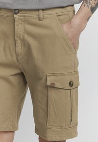 Shorts cargo beiges avec poches latérales, fabriqués en tissu résistant. Dotés d'une fermeture à bouton, de passants de ceinture et de détails de couture visibles.