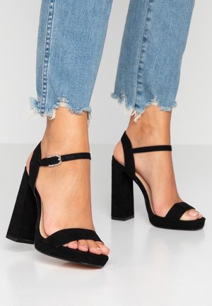 Fødder iført sorte ruskindssandaler med blokhæl og ankelremme, kombineret med lyseblå denimjeans med frynset kant på en hvid gulv.