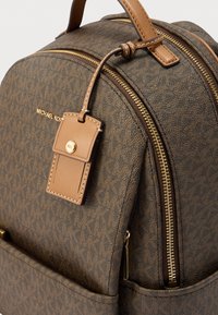 Sac à dos marron texturé avec des fermetures éclair dorées, des accents en cuir et une étiquette de bagage de marque attachée. Motif de logo distinctif sur toute la surface.