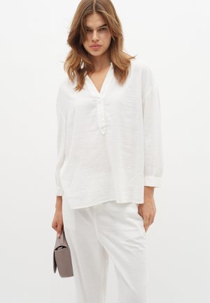 Blusa bianca in lino con scollo a V, vestibilità ampia e maniche lunghe. Presenta una texture sottile ed è abbinata a pantaloni bianchi sartoriali e a una borsa grigia.