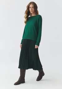 Maglione verde a maglia con orlo a coste, abbinato a una gonna plissettata di un verde scuro. Il modello indossa stivali marroni al ginocchio. Stile minimalista, colori solidi.