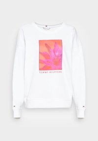 Sweatshirt blanc à manches longues, présentant un motif de fleur rose et le texte "TOMMY HILFIGER" en dessous. Matière en coton doux. Encolure ras-du-cou.