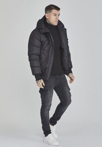 Schwarze Steppjacke mit Kapuze, Reißverschluss und elastischen Bündchen, kombiniert mit einem schwarzen Rollkragenpullover und dunklen Cargo-Jeans, abgerundet mit weißen Sneakers.