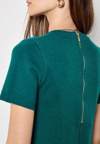 Top a maglia verde acqua con maniche corte, tessuto testurizzato e zip dorata visibile sul retro.