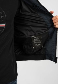 Giubbotto nero con un interno liscio e lucido; presenta un'etichetta Geox, chiusura con zip e orlo a costine. Il materiale sembra leggero e traspirante.