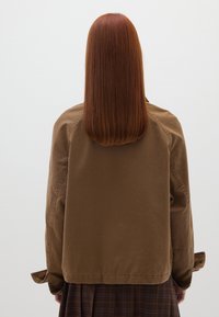 Braune Baumwolljacke mit lässiger Passform, sichtbaren Nähten und Knopfdetails an den Ärmelabschlüssen, von hinten vor einem schlichten Hintergrund gezeigt.