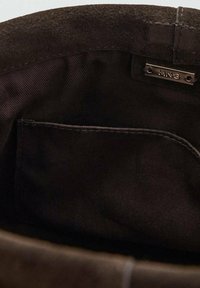 Interior de bolsa de ante en marrón oscuro con forro de algodón. Presenta un bolsillo sutilmente cosido y una pequeña etiqueta metálica con la inscripción "MNG."