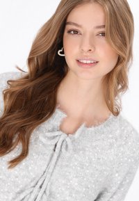 Szürke bolyhos pulóver flitterekkel, kerek nyakkivágással és elől megkötős részlettel. A modellen hosszú, hullámos haj van, szív alakú fülbevalót visel.