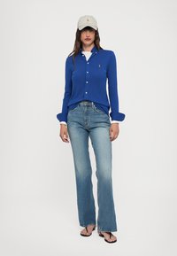 Blå långärmad skjorta med knappstängning, vita krageaccent, kombinerad med ljusblå utsvängda jeans och en ljusbeige basebollkeps.
