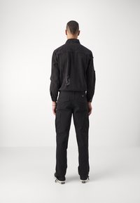 Fekete farmerdzseki hasítékokkal, gombos gallérral és hosszú ujjakkal, fekete cargo nadrággal párosítva, amely oldalzsebbel és slim fit szabással rendelkezik.