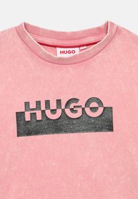 Rozā kokvilnas t-krekls ar noapaļotu kakla izgriezumu. Priekšpusē ir melns, spīdīgs "HUGO" laukums uz teksturēta fona.