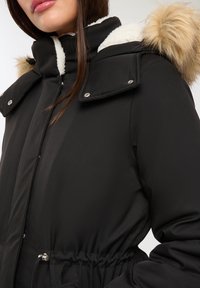 Czarna zimowa kurtka z kapturem wykończonym sztucznym futrem, zapinana na zamek błyskawiczny z przodu i z regulowanym pasem na sznurki. Posiada srebrne elementy metalowe i gładką teksturę materiału.