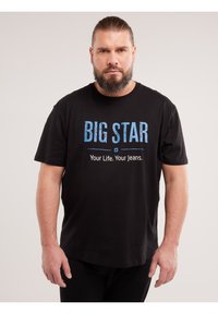 Odrasli muškarac s bradom nosi crnu majicu s plavim tekstom "BIG STAR" i bijelim tekstom "Your Life. Your Jeans." na jednobojnoj pozadini.